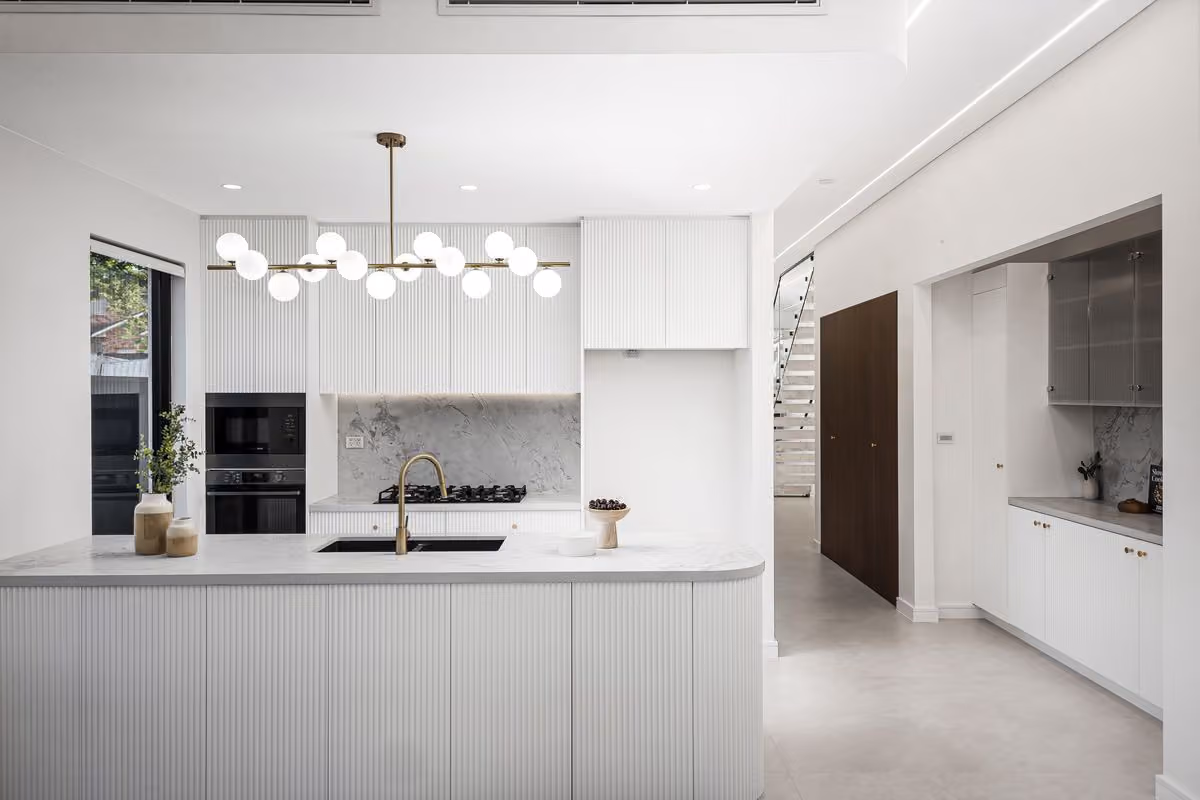 White kitchen with a long island, globe pendant lights, black appliances and a timber-lined pantry nook