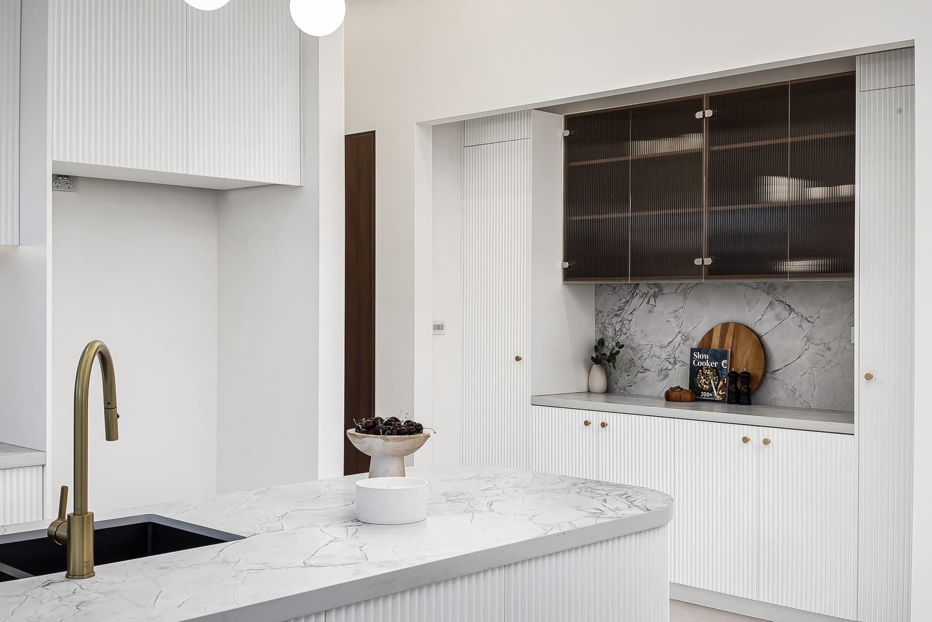 White stone benchtop with a brushed brass tap, fluted cabinetry and a dark glass cabinet behind