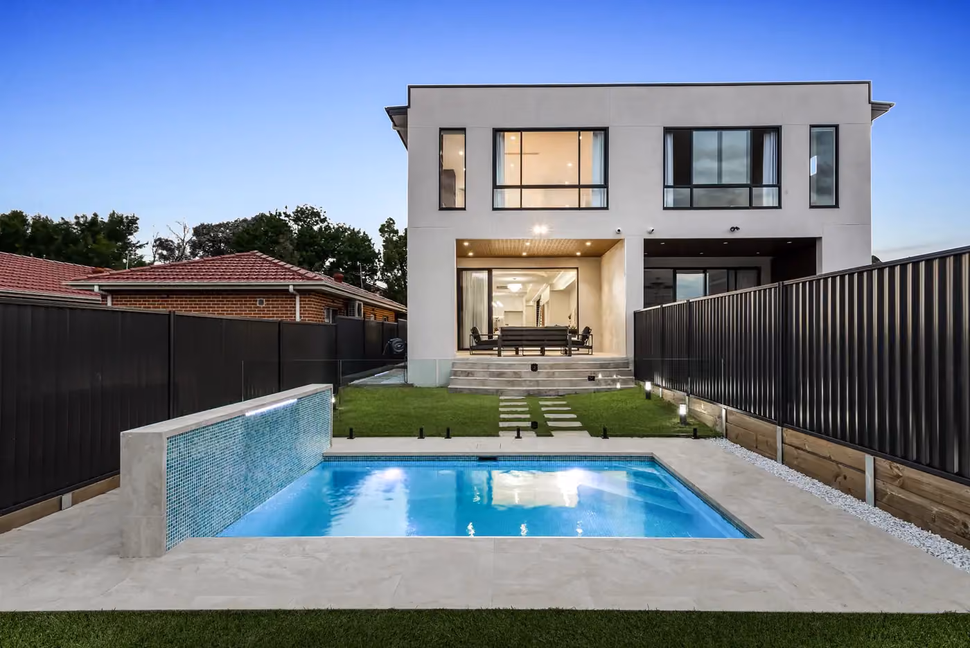 Rear of a two-storey family home at dusk with glowing windows, a pool and a stretch of lawn